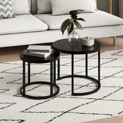 Two black round coffee tables are placed on a white rug with a black geometric pattern. A sofa is visible in the background.