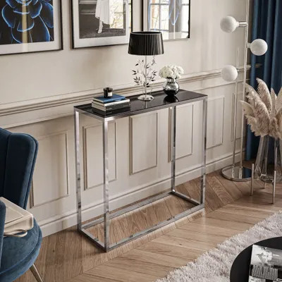 A modern console table with a chrome frame and black glass top, decorated with a lamp, books, and flowers, placed in a living room setting.