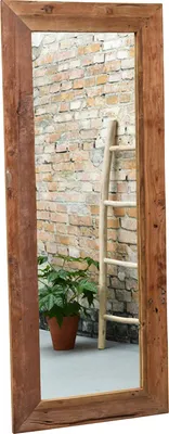 A tall, narrow mirror with a rustic wooden frame stands against a brick wall. A wooden ladder leans against the wall behind the mirror, and a potted plant sits on the floor in front of the mirror.