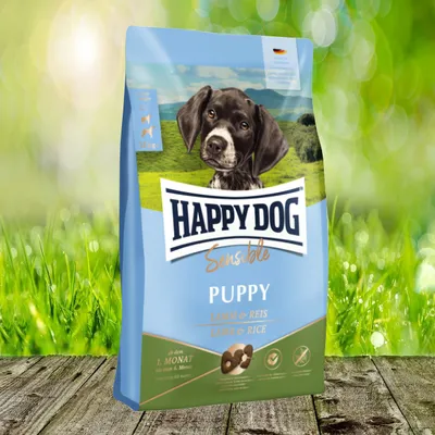A bag of Happy Dog Sensible Puppy dog food is displayed on a wooden surface with a blurred green grassy background and sunlight.