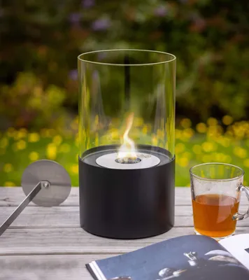 A bioethanol table fireplace is shown on a wooden table outdoors, with a cup of tea and a book nearby.
