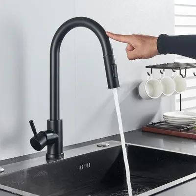 A black kitchen faucet with a pull-down sprayer is shown in use, with water flowing into a sink. A hand is touching the faucet, likely activating a sensor. Several white mugs are hanging on a rack in the background.
