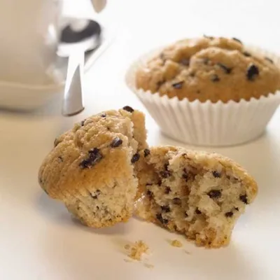 A close-up of a chocolate chip muffin, broken in half, with another muffin in the background and a partial view of a white mug and spoon.