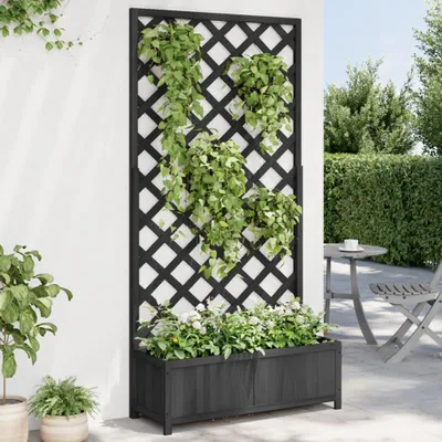 A planter box with a trellis attached, filled with plants, is placed against a white wall outdoors. A table and chair are visible in the background.