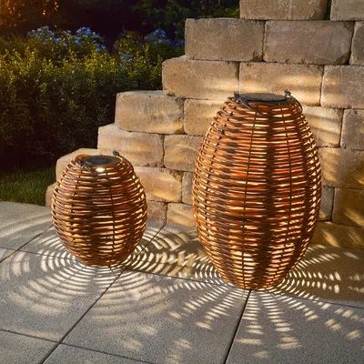 Two rattan solar lanterns are placed on a paved patio next to a stone wall at dusk, casting intricate shadows.