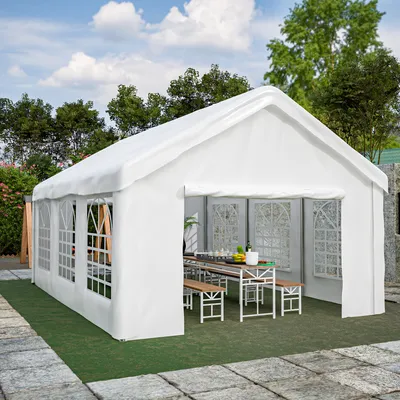 A white party tent set up outdoors with tables and benches inside, surrounded by greenery and a stone patio.