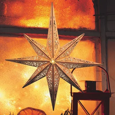 A wooden star ornament with intricate cutouts is illuminated from within, set against a warm, glowing background resembling a window with firelight. A red lantern is visible in the foreground.