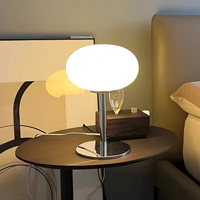 A minimalist glass table lamp sits on a wooden side table next to a bed.