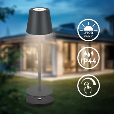 A dark gray portable table lamp is shown in a garden setting at dusk, with a house in the background.