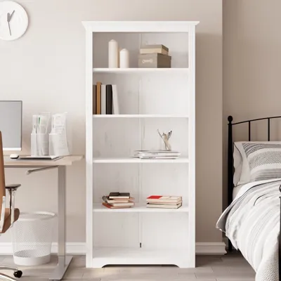 A white bookshelf filled with books and decorative items, placed in a room with a desk and a bed.