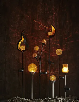 A collection of solar-powered garden lights with flame, crescent moon, and orb shapes, set against a dark, textured wooden background and a bed of small stones.