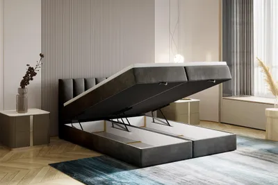 A dark gray boxspring bed with its storage compartments open, revealing the interior. The bed is in a modern bedroom setting with a nightstand and decorative elements.