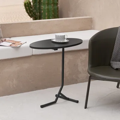 A black oval side table with a cup and saucer on it, next to a dark green chair and a cactus.