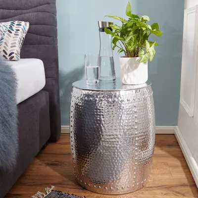 A silver hammered metal side table with a plant, a glass of water, and a carafe of water on top, next to a bed.