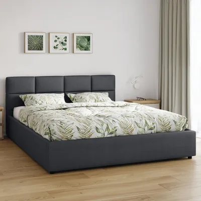 A dark gray upholstered bed with a botanical print duvet and pillows, set against a white wall with framed botanical prints and a light beige curtain.