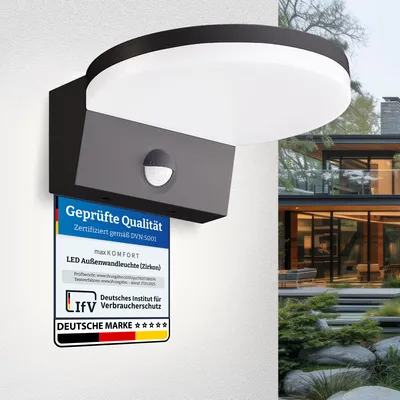 A black LED outdoor wall light with a motion sensor is mounted on a white wall, with a modern house and garden in the background. A certification label is attached to the light.