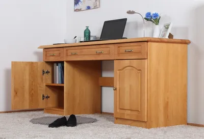 A wooden desk with drawers and cabinets, set up in a room with a white wall and a plush carpet. A laptop, lamp, and decorative items are on the desk surface.
