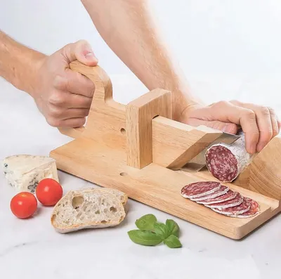A person is using a wooden slicer to cut salami, with bread, cheese, tomatoes, and basil on the side.