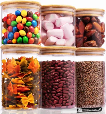 Sechs klare Glas-Lebensmittelaufbewahrungsbehälter mit Bambusdeckeln, gefüllt mit verschiedenen Trockenwaren wie Bonbons, Marshmallows, Datteln, Nudeln, Bohnen und Linsen, mit einem weißen Kreidestift im Vordergrund