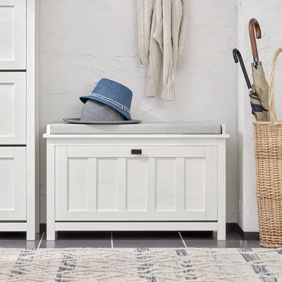 A white shoe bench with a gray cushion is placed in an entryway, with two hats on top and a sweater hanging on the wall behind it. An umbrella stand with umbrellas is to the right.
