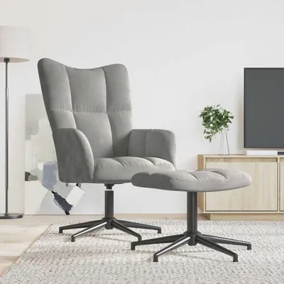 A light gray velvet armchair and ottoman are placed on a rug in a living room setting. A floor lamp is visible on the left, and a TV stand with a plant and television is on the right.