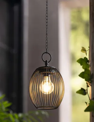A black metal solar LED lantern with a visible light bulb hangs from a chain outdoors, with greenery visible on the right.