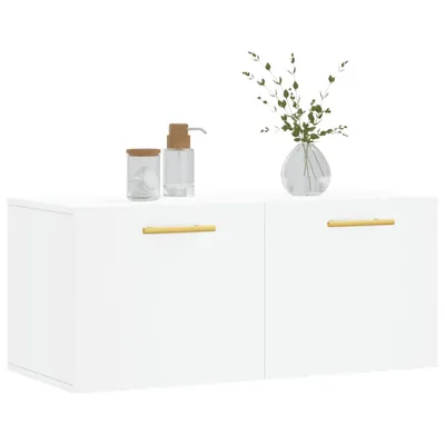 A white wall-mounted cabinet with two doors, each featuring a gold handle. On top of the cabinet are two small containers and a vase with greenery.