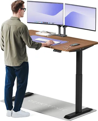 A man stands at a standing desk with two monitors, a keyboard, and a mouse.