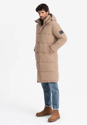 A man wearing a long beige puffer jacket and blue jeans, standing against a white background.