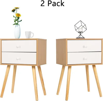 Two identical bedside tables with wooden legs and white drawers are shown. One table has a vase with yellow flowers and a white mug on top. The other has a geometric decorative item with a candle.