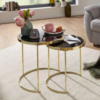 A set of nesting tables with black glass tops and gold metal frames, styled with a vase of flowers, a teacup, and a muffin.