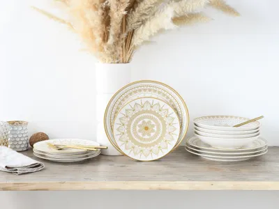 A set of white and gold mandala-patterned plates and bowls are arranged on a wooden shelf, with a vase of dried pampas grass in the background.