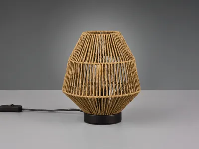 A table lamp with a woven rattan-like shade sits on a plain surface against a grey background.