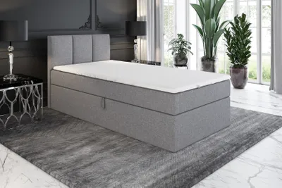 A grey boxspring bed with a mattress and topper is shown in a bedroom setting.