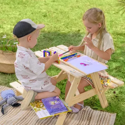 Two children are drawing and playing at a wooden picnic table outdoors on a blanket.
