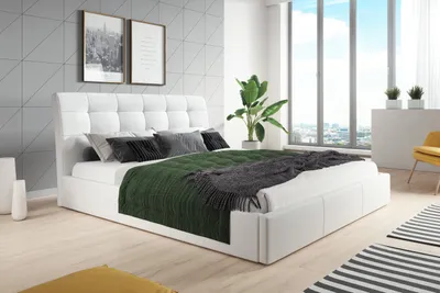 A modern bedroom with a white upholstered bed, green and gray blankets, and a large window overlooking a city.