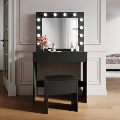 A black vanity table with a lighted mirror and a matching stool in a room with decorative wall molding and wooden flooring.