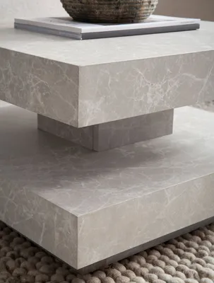 A close-up view of a modern, square coffee table with a marble-like finish, resting on a textured rug.