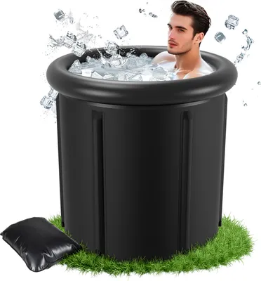 A man is sitting in a black inflatable ice bath filled with ice and water, with ice cubes splashing around him. The bath is placed on a patch of green grass.