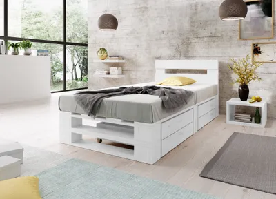 A bedroom scene with a white wooden bed frame, grey bedding, and a yellow pillow. The room has a concrete wall and a large window with a view of trees.