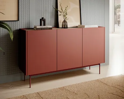A modern red sideboard with three doors, standing on thin metal legs, is placed against a textured gray wall. Books and a vase with branches are on top of the sideboard. A rug is on the floor in front of it.
