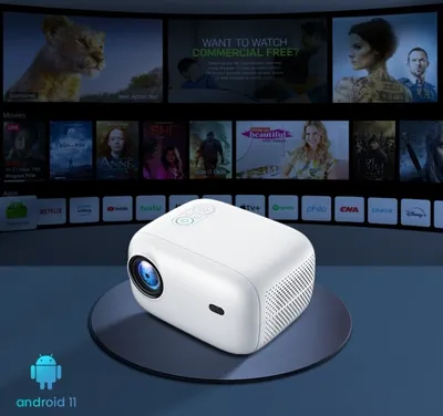 A white projector is placed on a dark circular stand in front of a large screen displaying various movie posters and app icons.