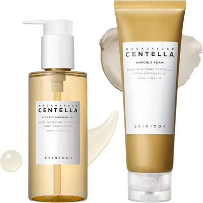 Two skincare products, a cleansing oil and a foam cleanser, are displayed against a white background with subtle cream-colored smudges.