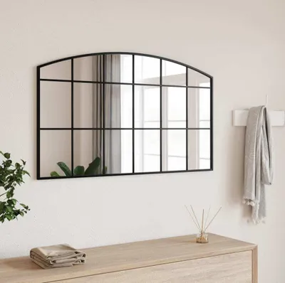 A black arched mirror with a grid design hangs on a white wall above a wooden dresser. A gray scarf is draped over a hook to the right of the mirror, and a small plant and diffuser are on the dresser.