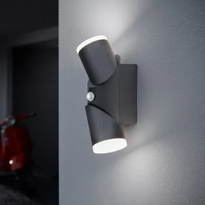 A dark gray outdoor LED wall light with two adjustable heads and a motion sensor is mounted on a textured gray wall. To the left, a red vintage scooter is partially visible in a dimly lit entryway.