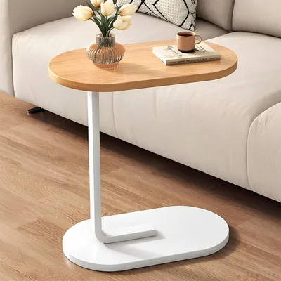 A small oval-shaped side table with a wooden top and a white base and leg, placed next to a cream-colored sofa. A vase with flowers and a book with a cup on top are on the table.