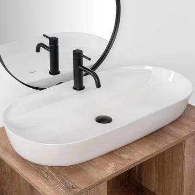 A white oval countertop sink with a black faucet is shown on a wooden vanity. A round mirror is visible in the background.