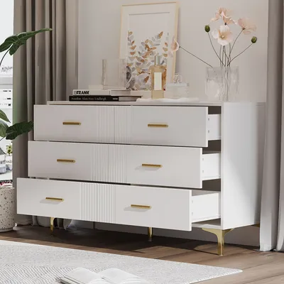 A white dresser with gold accents and drawers slightly open, placed in a room with curtains and a plant.