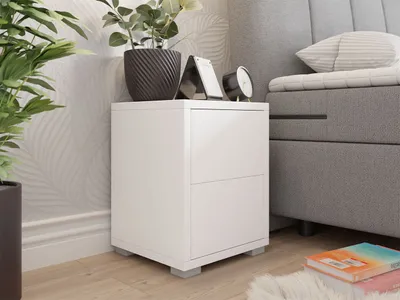 A white bedside table with two drawers is placed next to a bed. There are plants and a clock on top of the table.