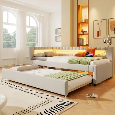 A bedroom scene featuring a beige upholstered daybed with a pull-out trundle bed, illuminated by LED lighting.